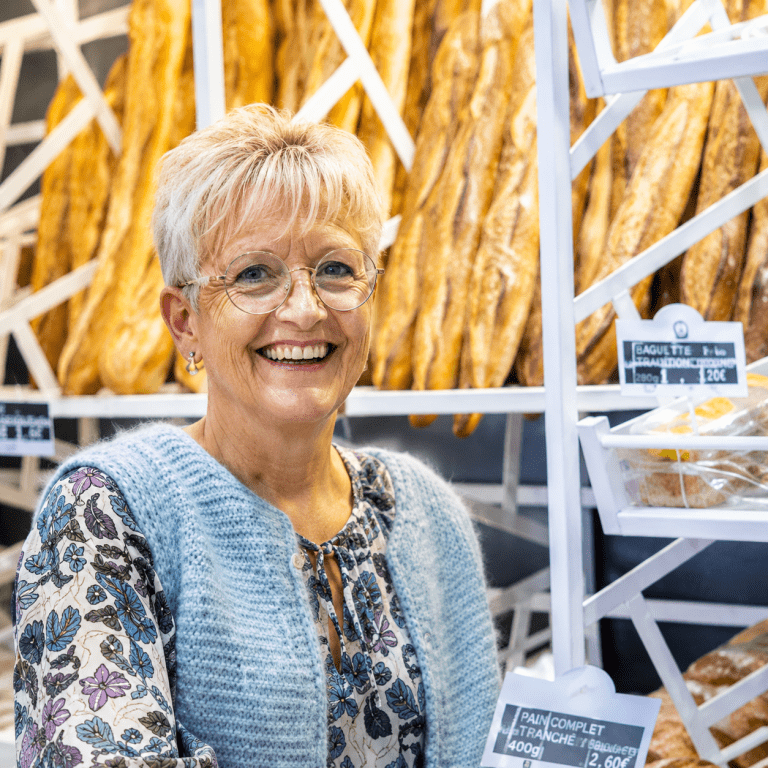 Boulangère souriante devant un présentoir de baguettes dans une boulangerie artisanale à Vitry-sur-Seine.