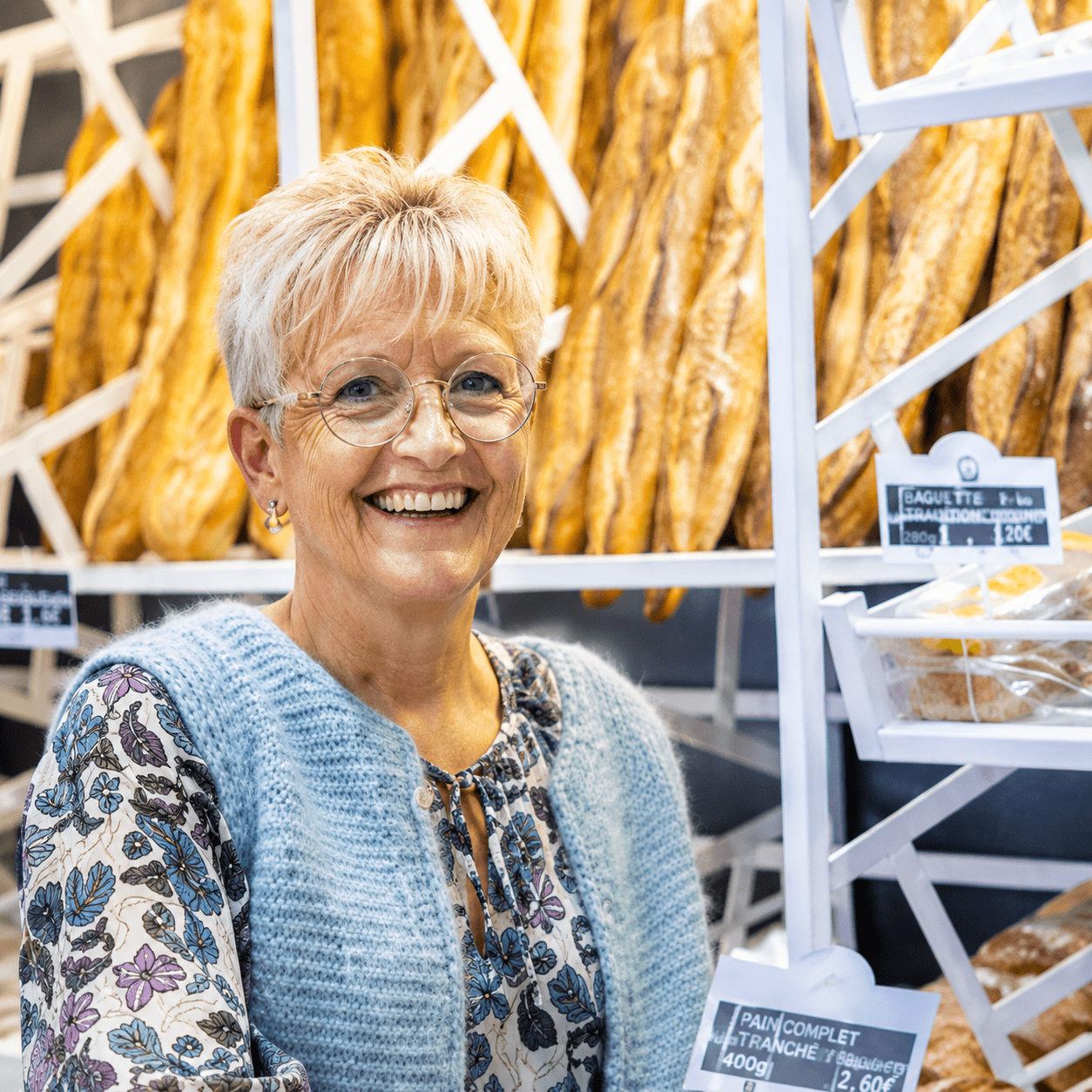 Boulangère souriante devant un présentoir de baguettes dans une boulangerie artisanale à Vitry-sur-Seine.