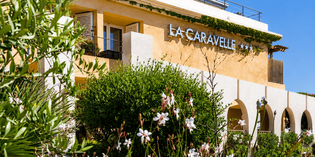 Façade de l'Hôtel La Caravelle à Calvi, en Haute-Corse, entourée d'un jardin méditerranéen luxuriant, à quelques pas de la plage et du centre historique.