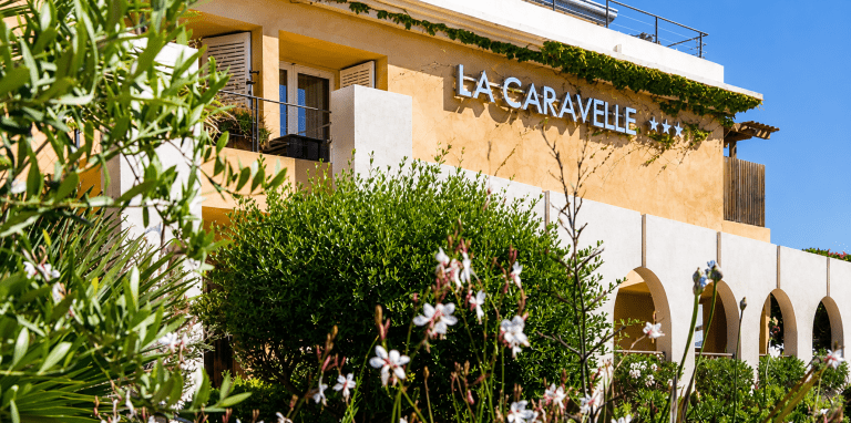 Façade de l'Hôtel La Caravelle à Calvi, en Haute-Corse, entourée d'un jardin méditerranéen luxuriant, à quelques pas de la plage et du centre historique.