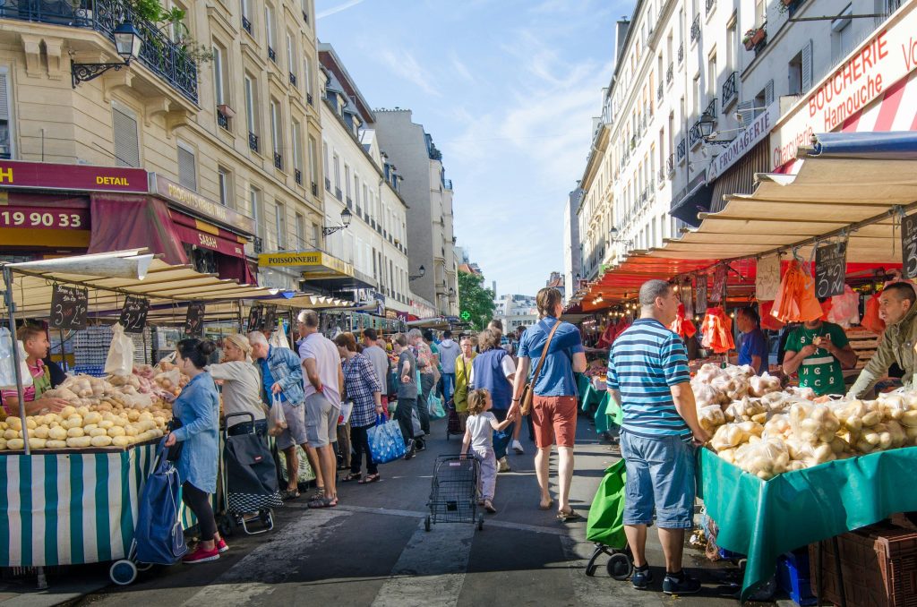 Marché de proximité avec des commerçants alimentaires et des clients dans une rue commerçante.