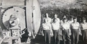 Photographie d’archives en noir et blanc montrant un groupe de jeunes femmes devant une machine industrielle pendant la Seconde Guerre mondiale.