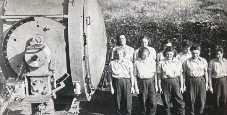 Photographie d’archives en noir et blanc montrant un groupe de jeunes femmes devant une machine industrielle pendant la Seconde Guerre mondiale.